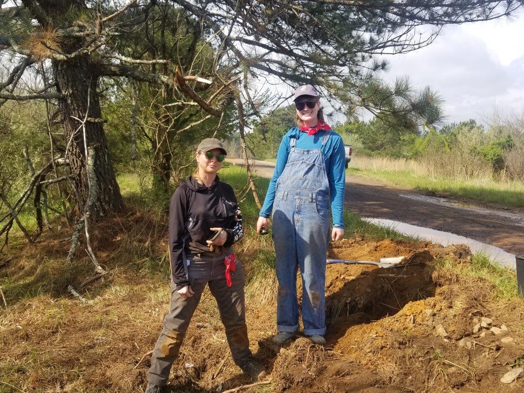 Two women stand on side of road near shovel.