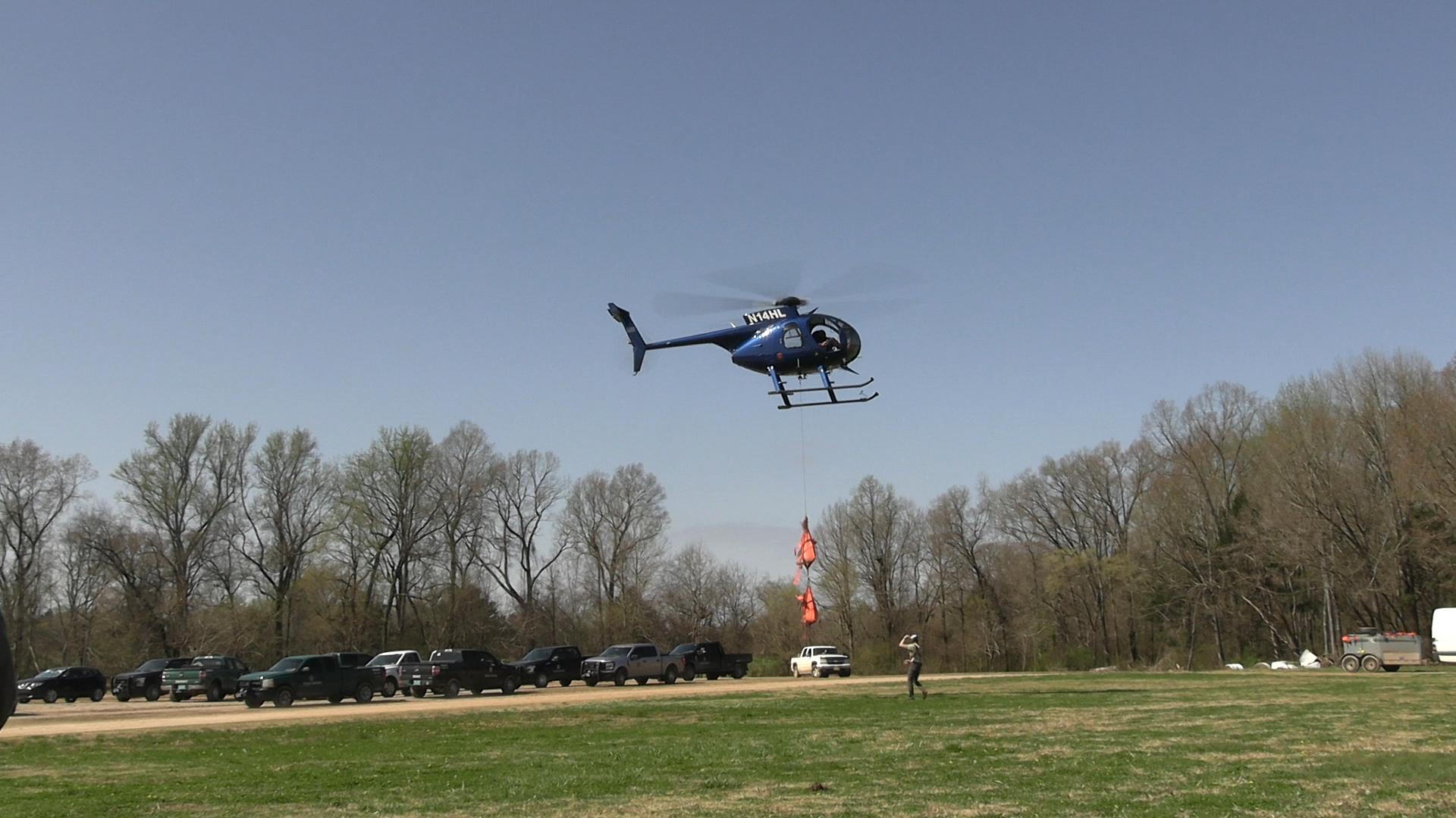 Helicopter carries two deer to man standing below on the ground.