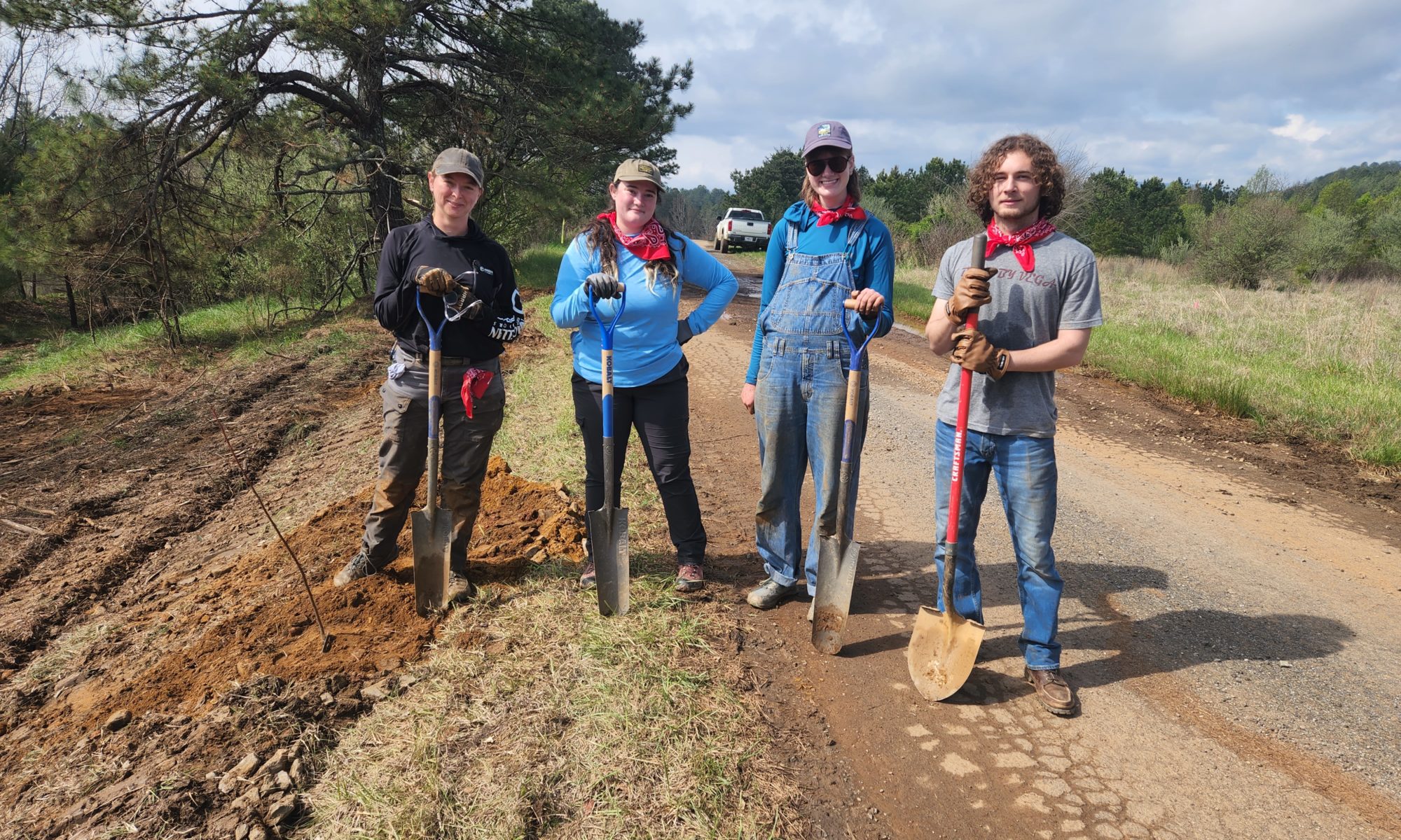 Four college students stand with shovels by road.