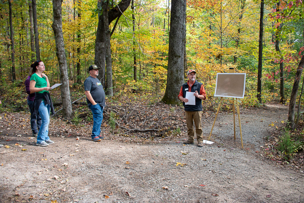 Man stands beside easel in a wooded area with people standing on his left.