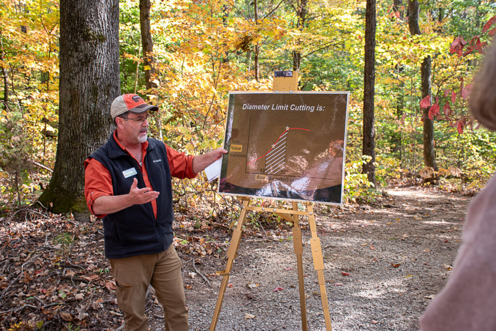 Man gestures to sign on easel in wooded area while person in foreground listens.