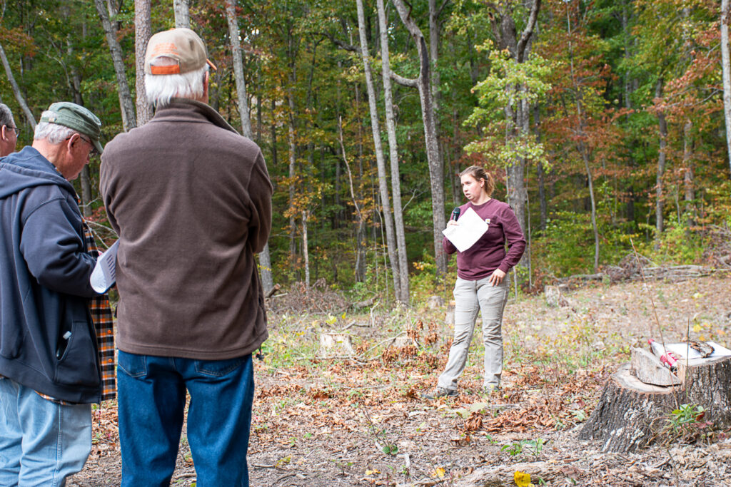 Woman speaks into microphone while people listen in a wooded area.