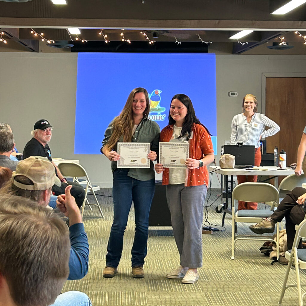 Two women hold awards while smiling at a group meeting.