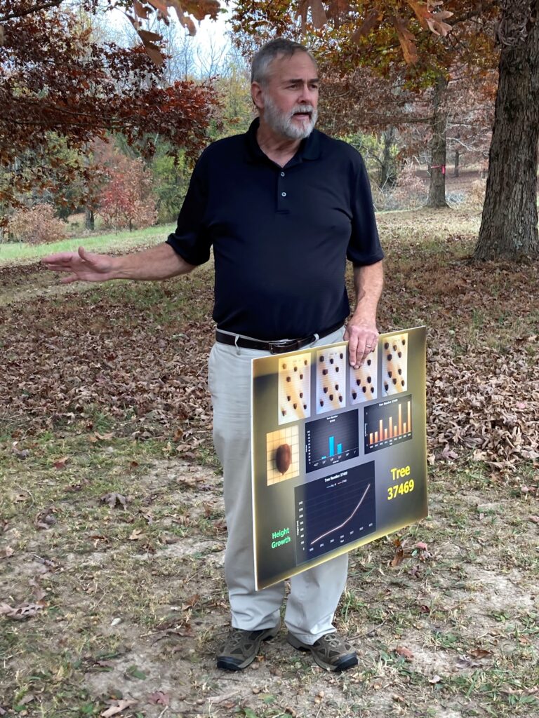 Man stands while talking and holding a sign outside surrounded by trees
