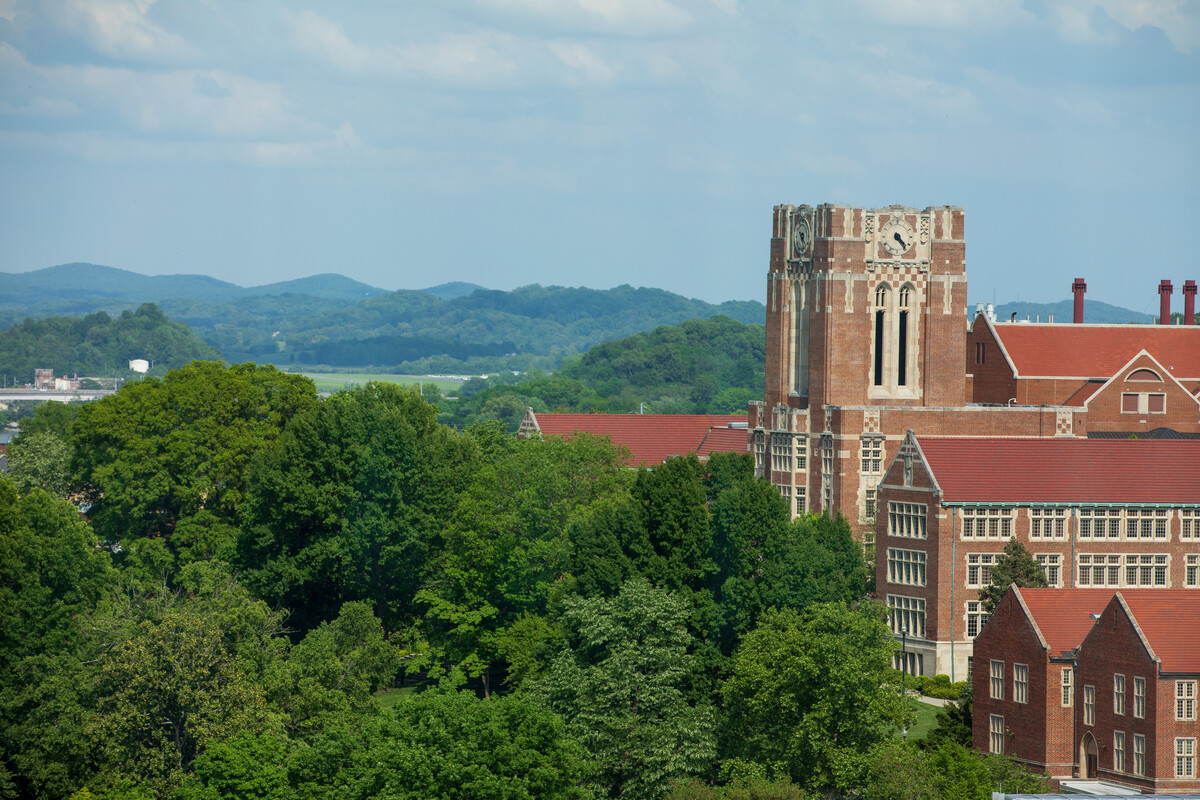 Aerial view shows Ayres Hall on the University of Tennessee campus with trees in the foreground and mountains in the background under a blue sky.