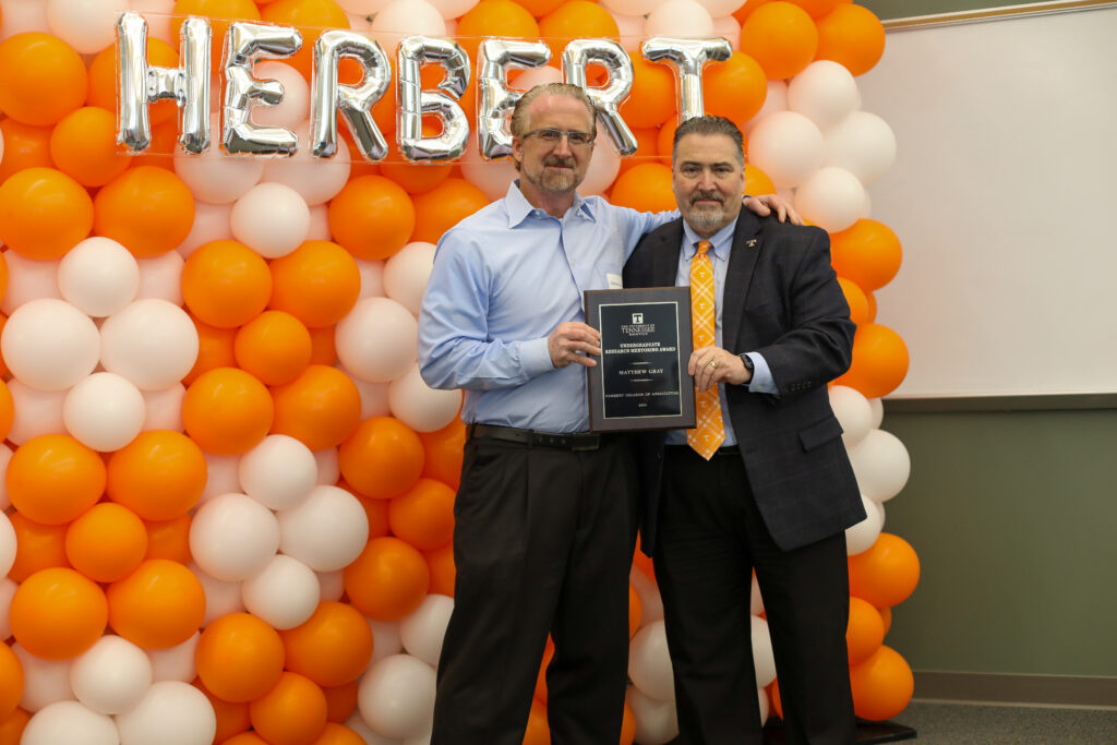 Two men stand in front of orange and white balloons, holding a plague.