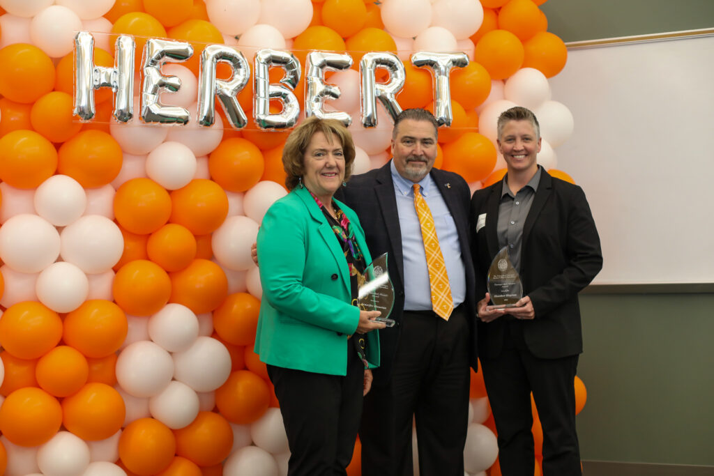 Three people stand in front of orange and white balloons. Two of them hold awards.