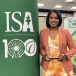 Woman in salmon jacket and orange and white dress poses with award next to International Society of Arboriculture sign.