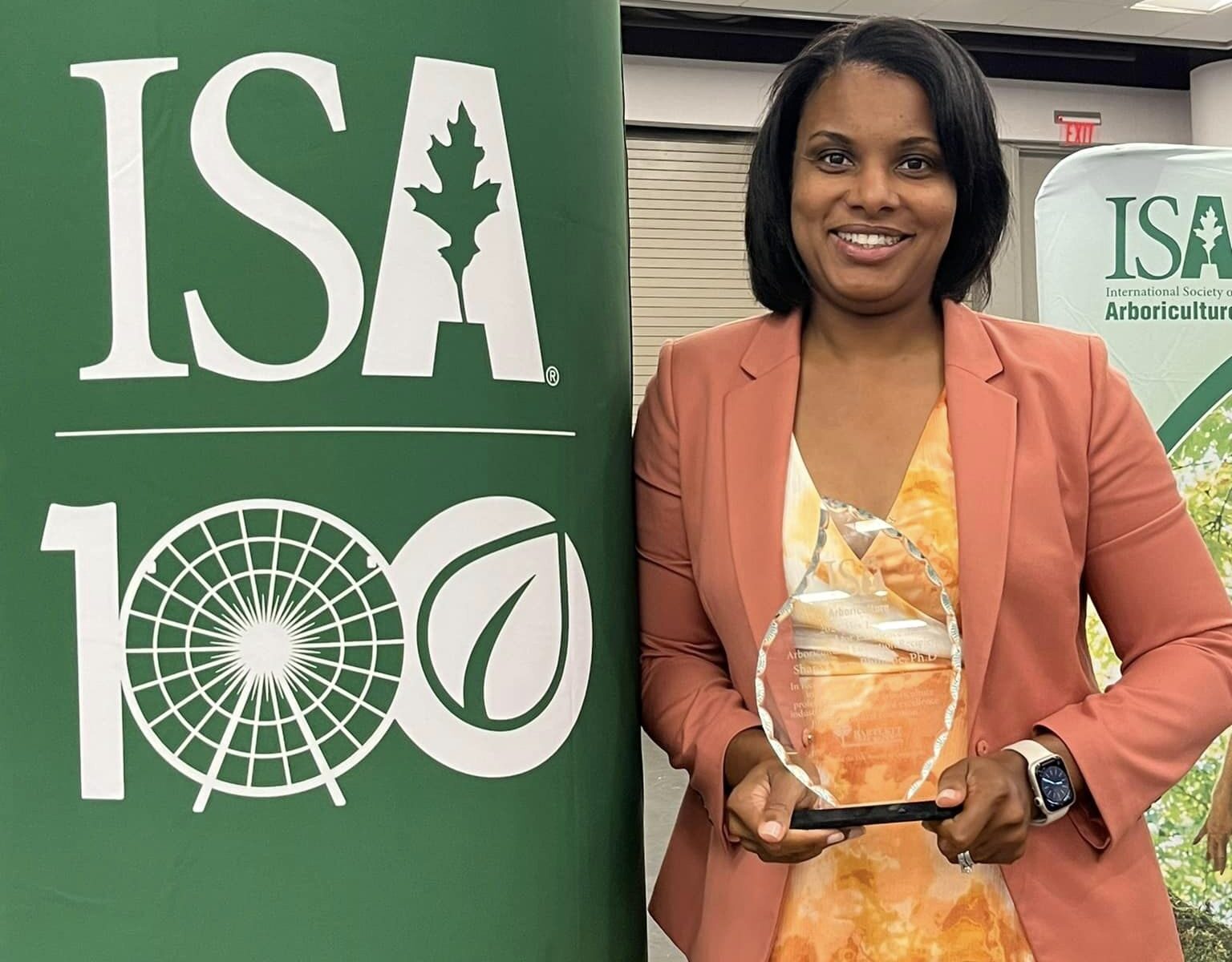 Woman in salmon jacket and orange and white dress poses with award next to International Society of Arboriculture sign.