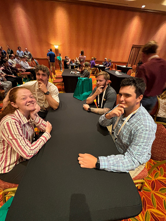 Four people sit at a table in a room full of other people.