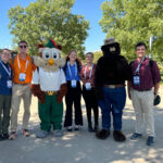 Five people pose with a bird mascot and the Smokey the bear mascot with trees behind them.