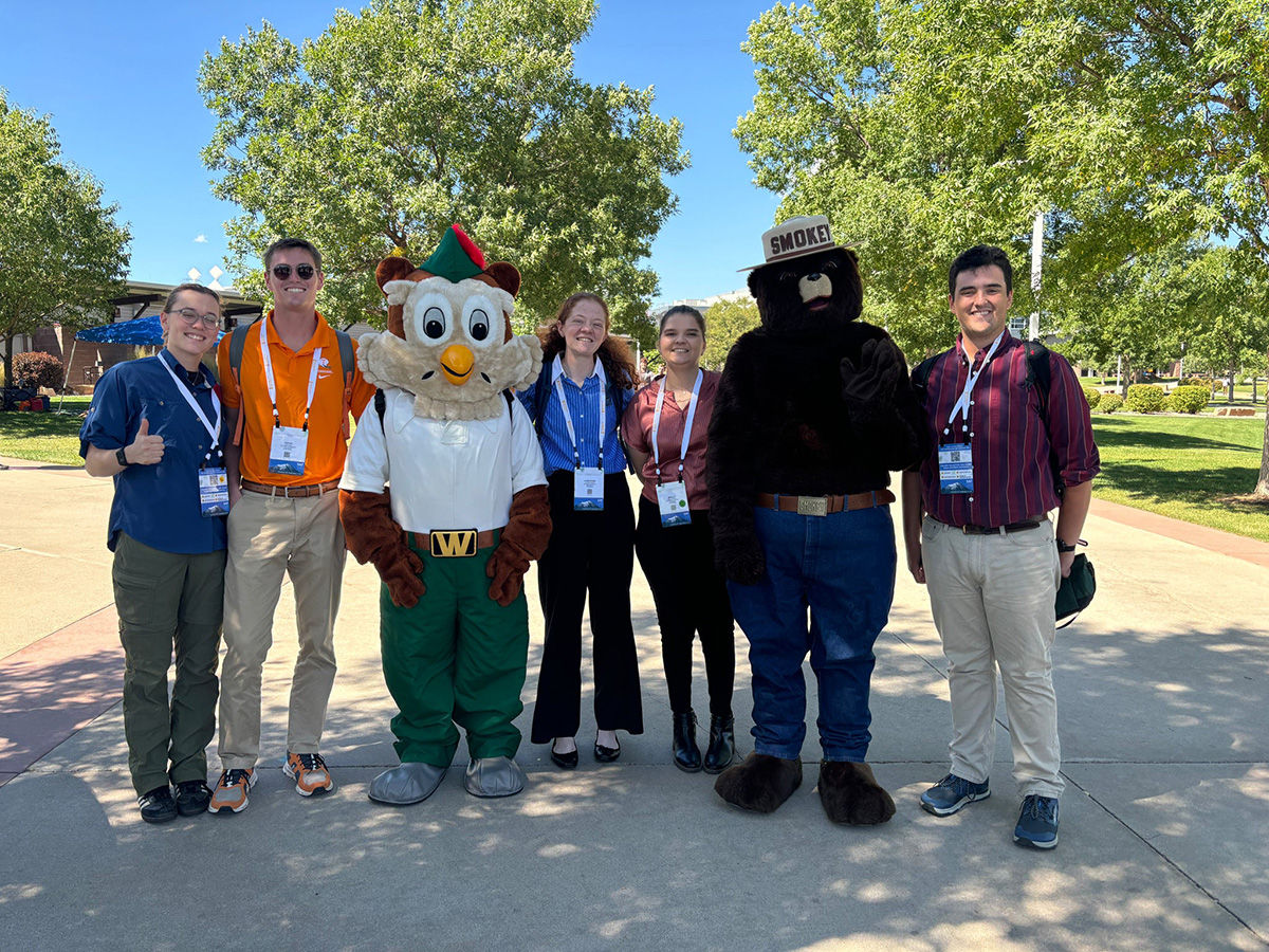 Five people pose with a bird mascot and the Smokey the bear mascot with trees behind them.