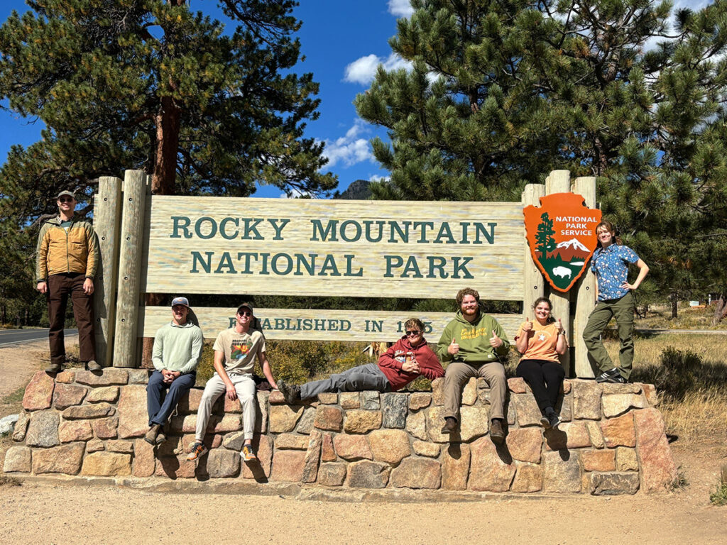 Seven people pose with the Rocky Mountain National Park sign.