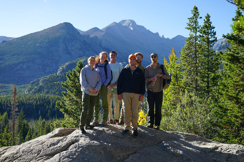 Six people stand and smile on a boulder with mountains and trees in the backgrounds.