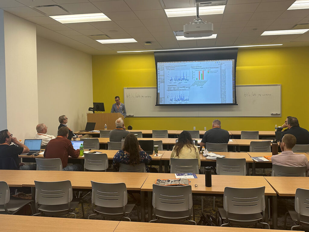 Eleven people sit at tables listening to a man giving a presentation in a room with a yellow wall.