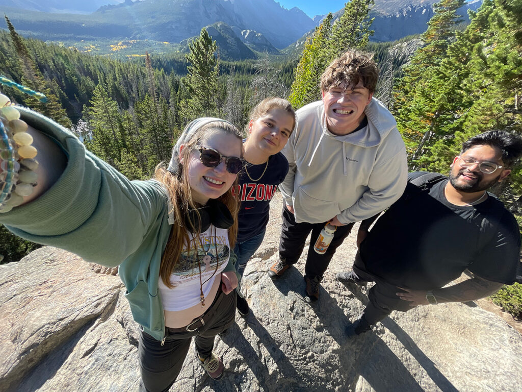 Four people pose on a boulder with trees and mountains in the background.