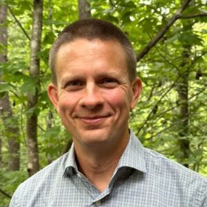 Man in button-up shirt smiles in front of a wooded area.