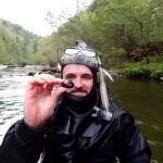 Man in wet suit and snorkel holds a mussel while standing in a creek.