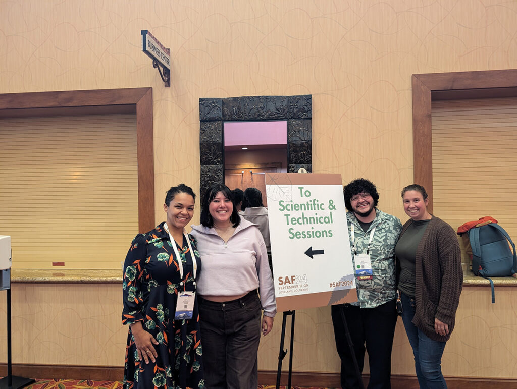 Four people stand in a room with a sign saying, "To Scientific and Technical Sessions."