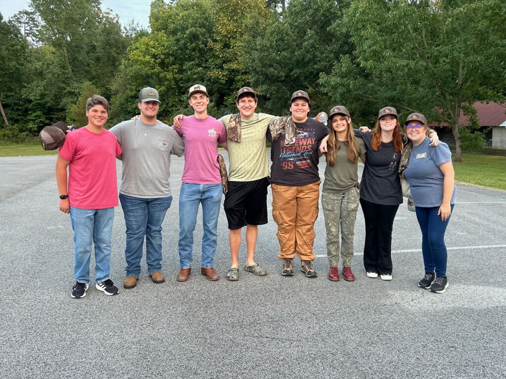 Eight people stand with their arms around each other in a parking lot with trees behind them.