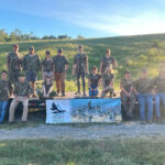 Fourteen people wearing camouflage sit or stand next to a trailer in front of a grassy hill.