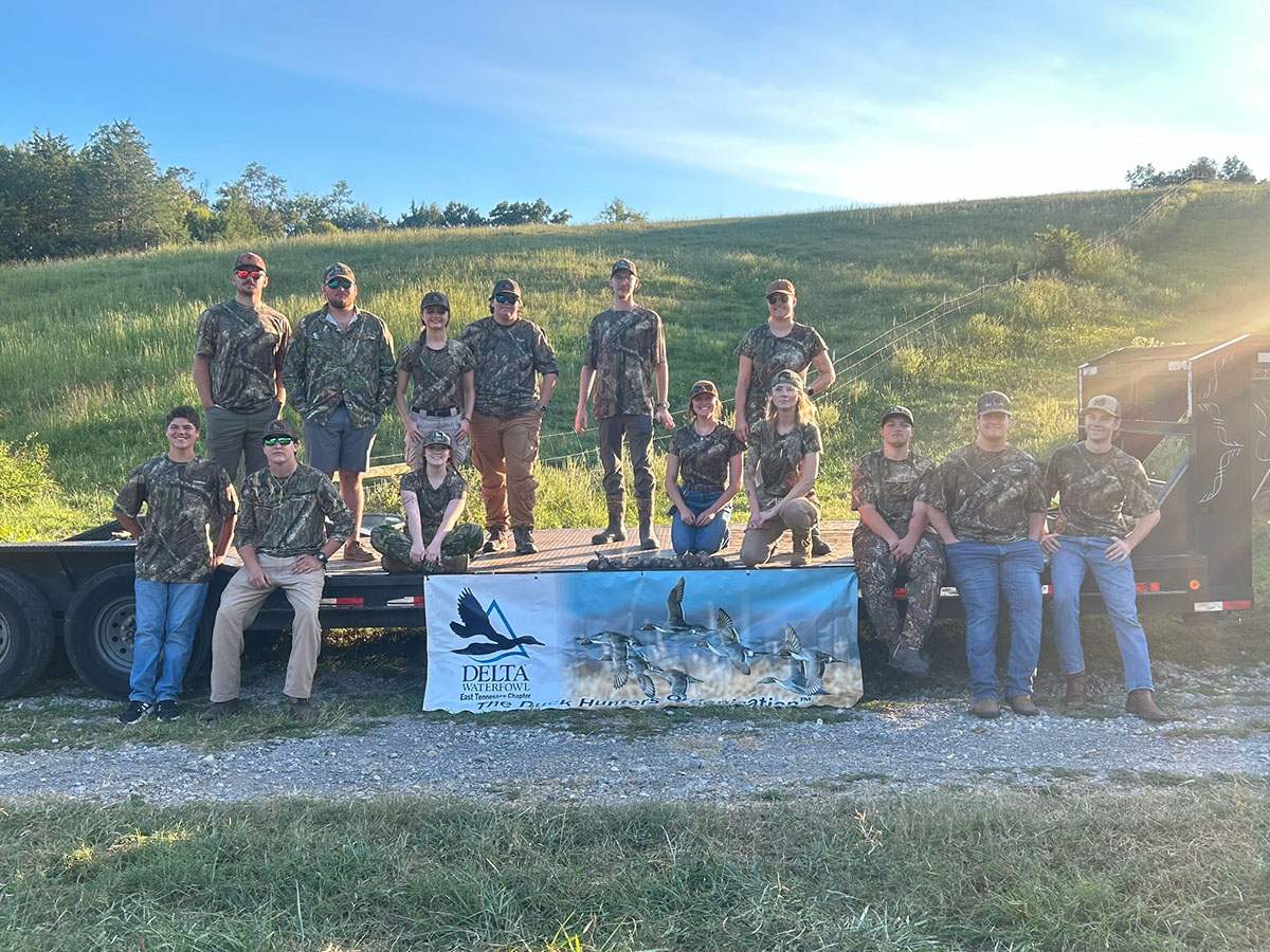 Fourteen people wearing camouflage sit or stand next to a trailer in front of a grassy hill.