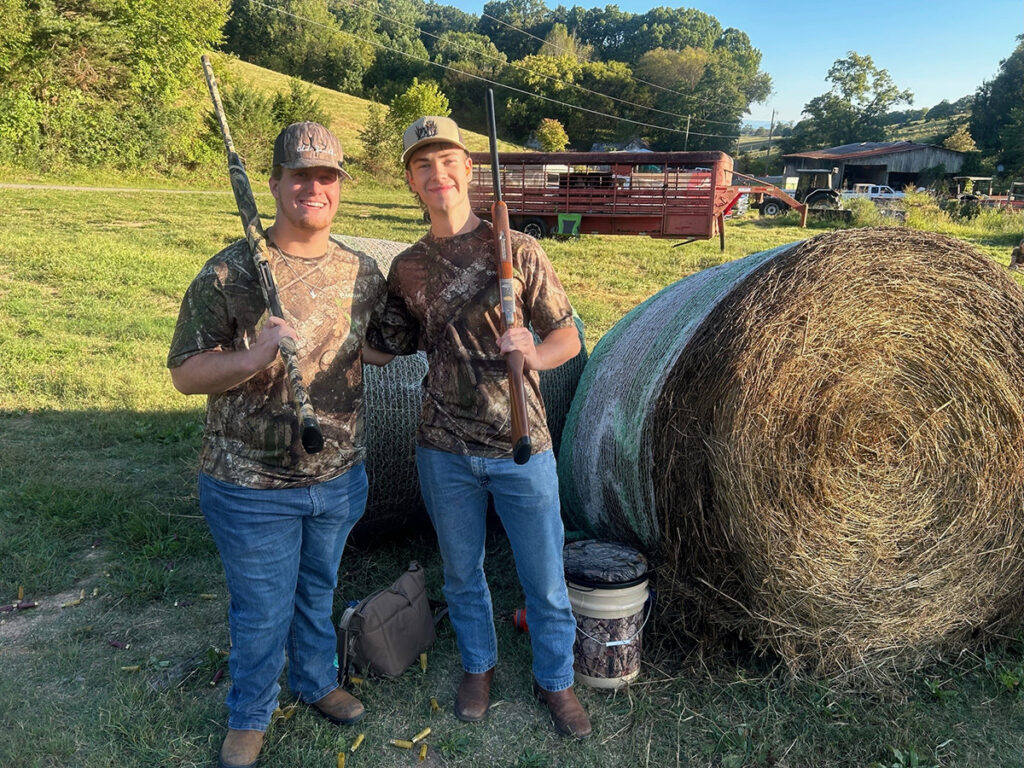 Two boys stand beside a hay bale with guns.