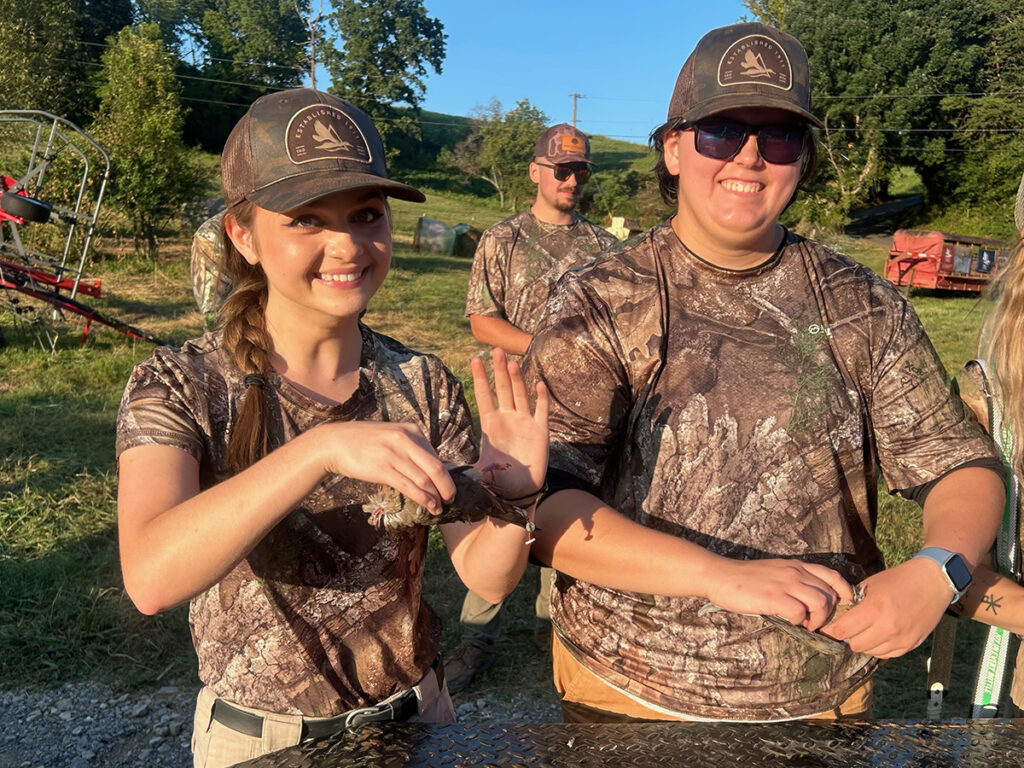 Two people wearing camouflage hold dead doves in their hands while out in a field.