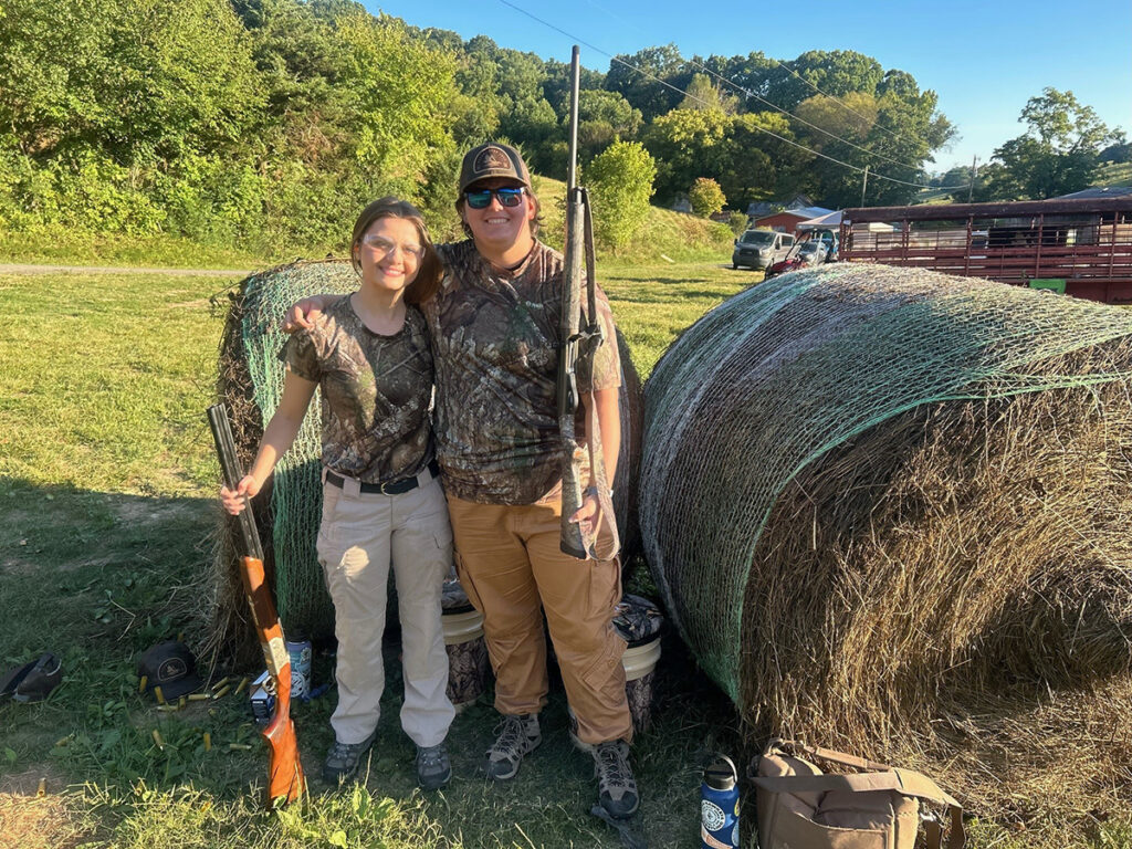 Two people stand with guns next to hay bales.