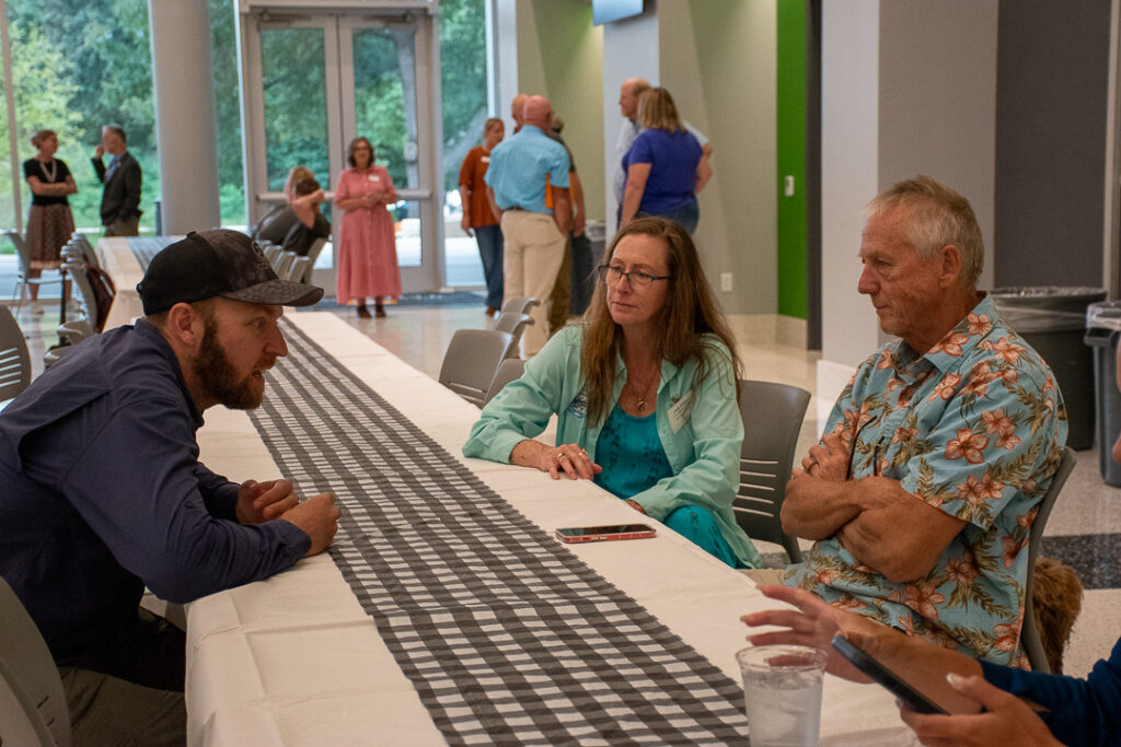 Three people sit at a table talking with others in the background.