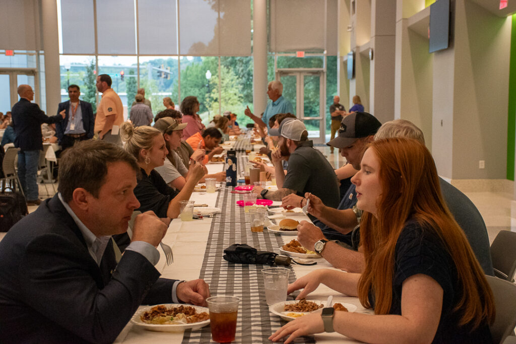 Several people sit at a long table while talking and eating.