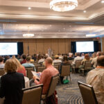 People sit in chairs and at tables listening to a speaker on a stage.