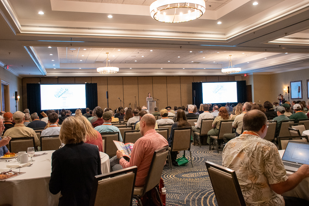 People sit in chairs and at tables listening to a speaker on a stage.