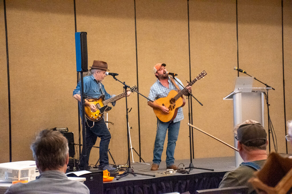 Two men play guitars on a stage in front of people.