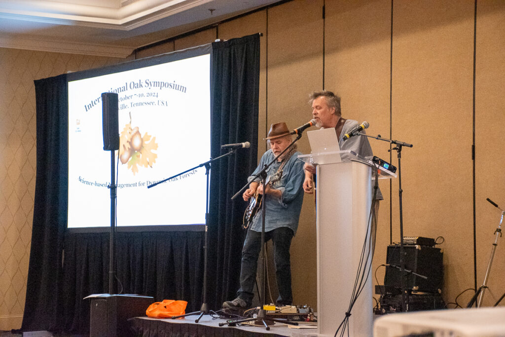 One man sings and another plays guitar on a stage beside a projector screen.