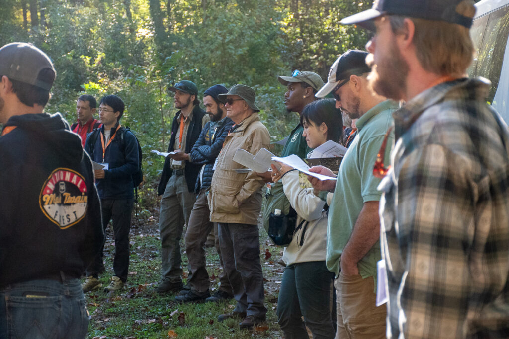 People stand around holding papers in a wooded area.