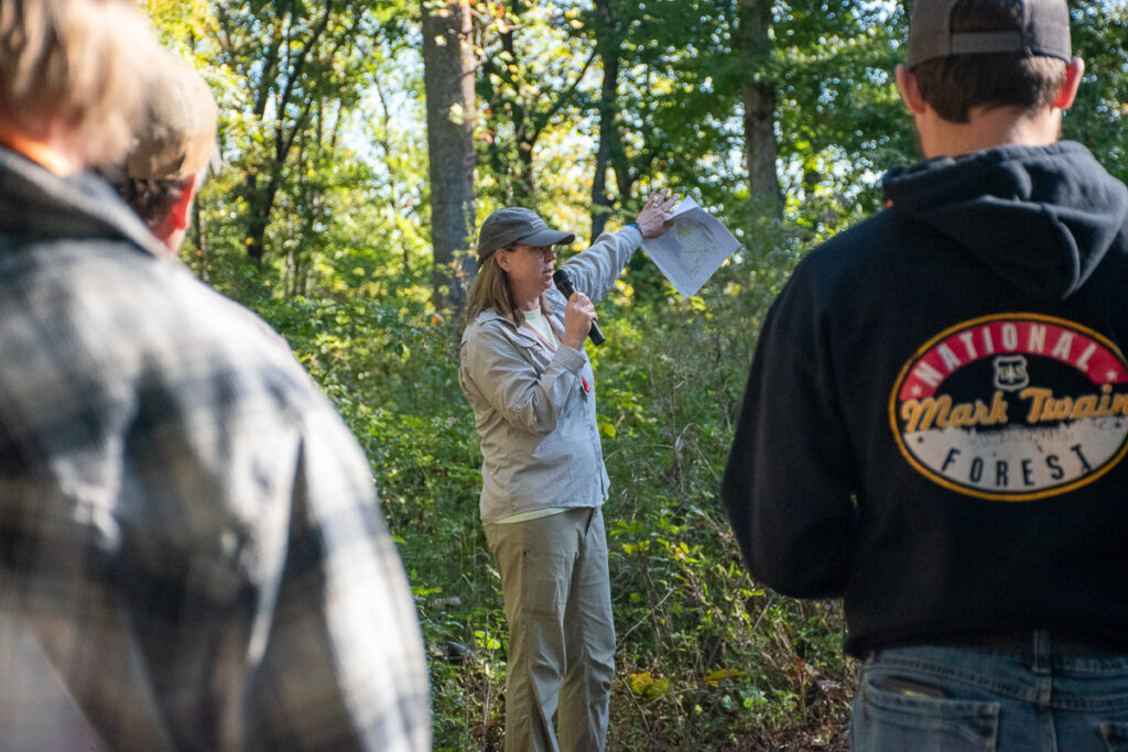 A woman speaks on a microphone to a group of people in a wooded area.