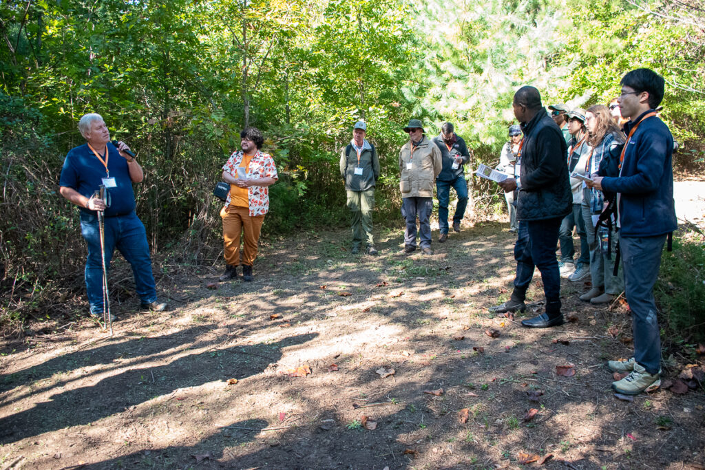 A man speaks to a group of people on a microphone while standing near trees.