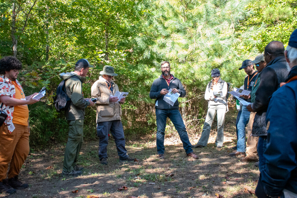 People stand and listen to a man speak near trees.