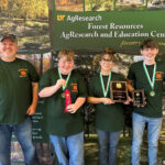 An adult man stands next to two boys and one girl at the Forest Resources AgResearch and Education Center with a sign behind them. The boys and girl wear medals and hold ribbons and plagues.