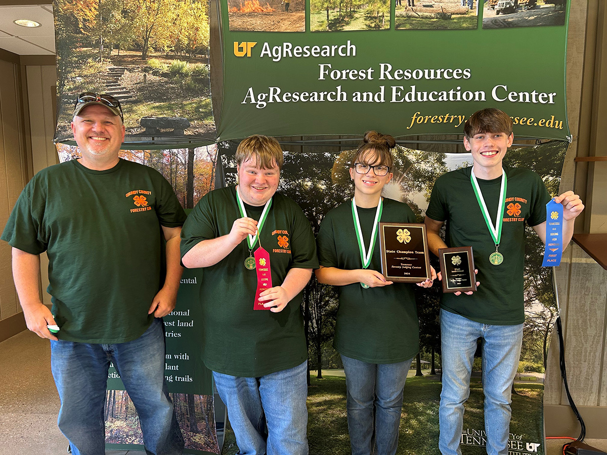 An adult man stands next to two boys and one girl at the Forest Resources AgResearch and Education Center with a sign behind them. The boys and girl wear medals and hold ribbons and plagues.