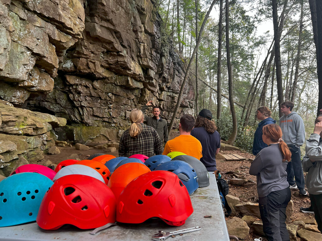 Seven people stand listening to an instructor speak in front of a rock wall with helmets of different colors in the foreground on a table with a wrench.