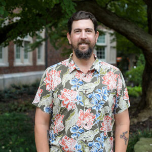 Man wearing floral button up shirt stands in front of building and tree.