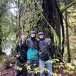 Three people stand with their arms around each other in front of a tree.