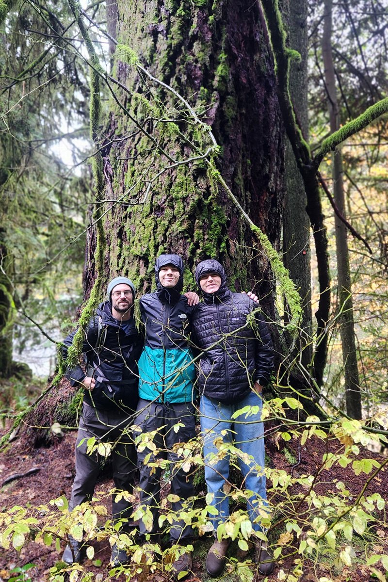 Three people stand with their arms around each other in front of a tree.