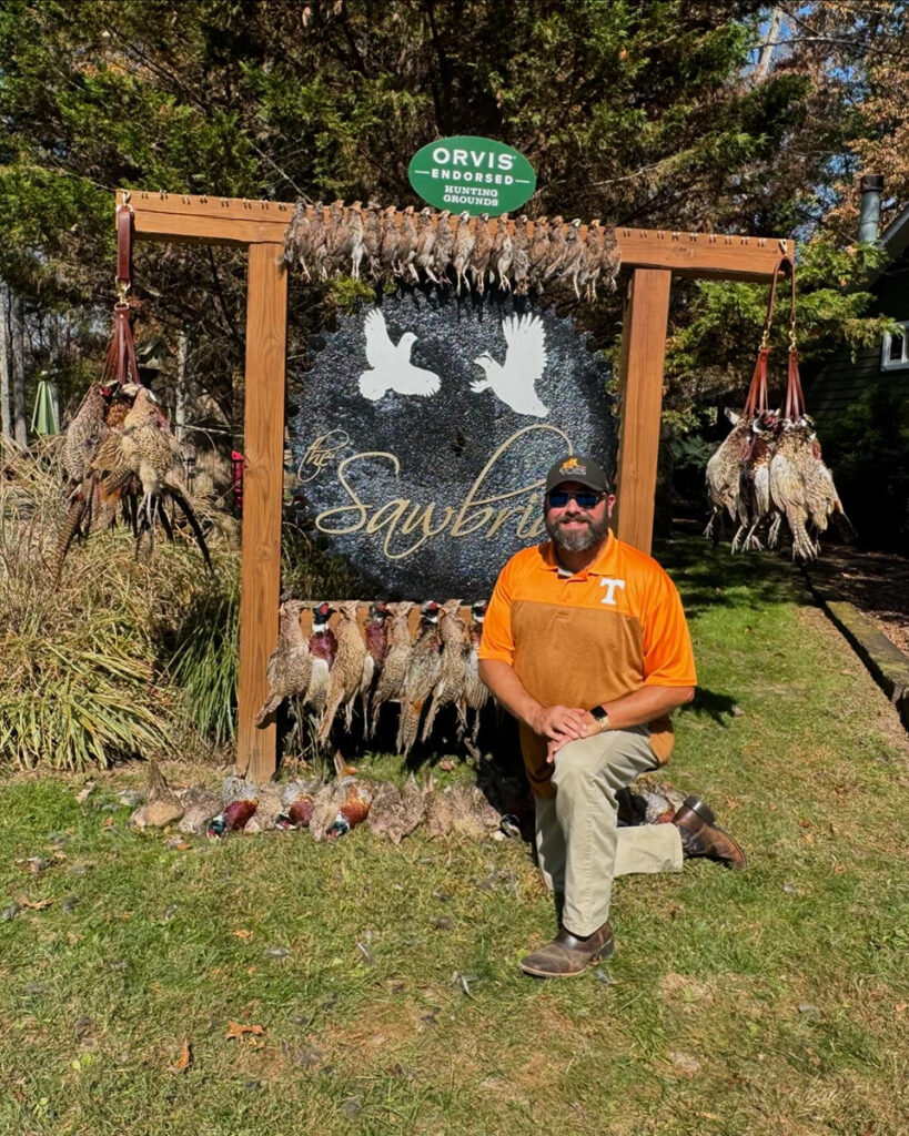 A man kneels on one knee in front of a sign with the words, "the Sawbriar," and dead quail hanging on both sides.