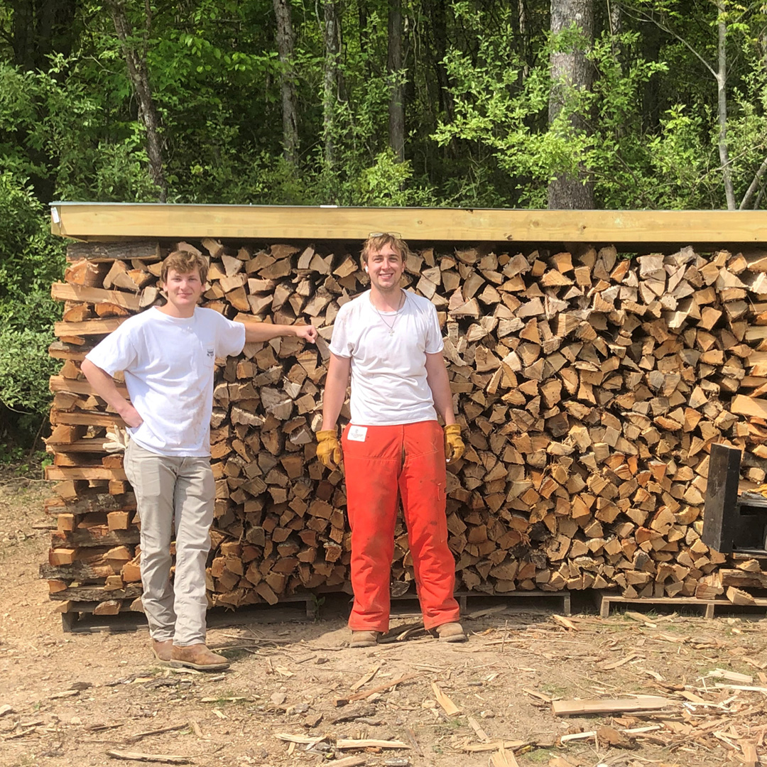 Two men stand in front of stockpile of firewood.