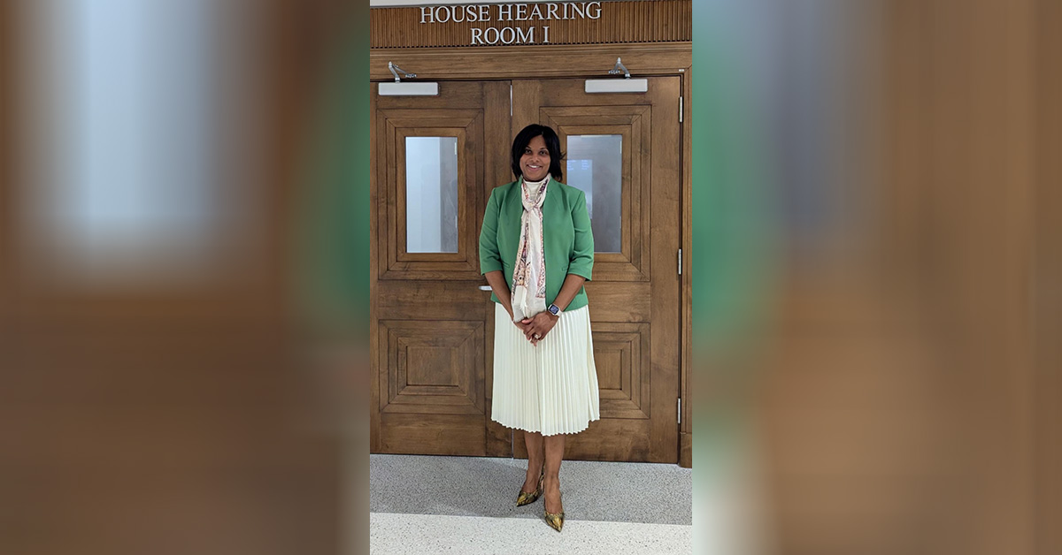 Woman in green jacket and cream skirt stands before double doors.