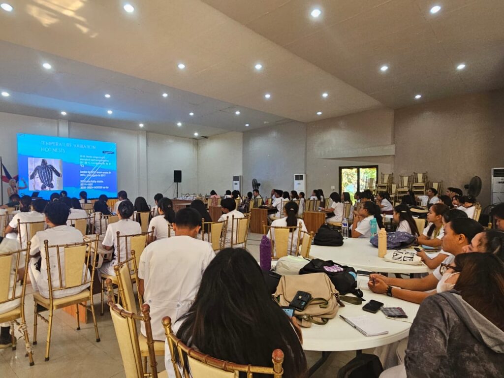 Several people sit at tables while a person gives a presentation at the front of a room.