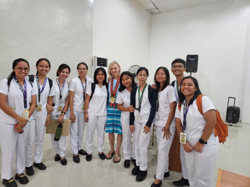 Several students in white uniforms pose for a photo with a women wearing a blue dress.